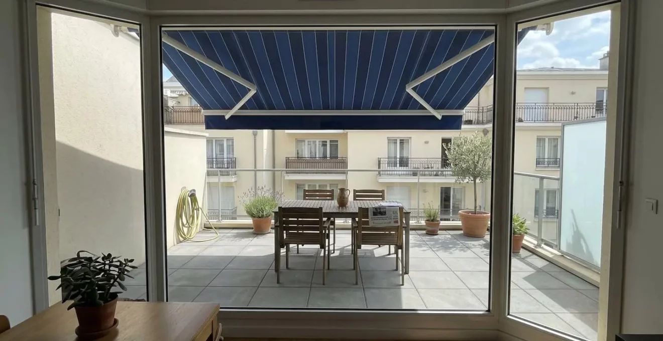 Terrasse française contemporaine vue depuis l'intérieur à travers une baie vitrée, store banne déployé projetant une ombre définie sur le mobilier de jardin et le dallage