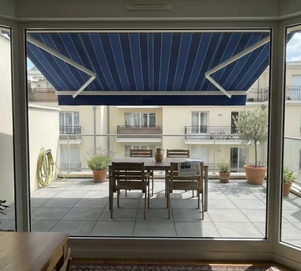 Terrasse française contemporaine vue depuis l'intérieur à travers une baie vitrée, store banne déployé projetant une ombre définie sur le mobilier de jardin et le dallage